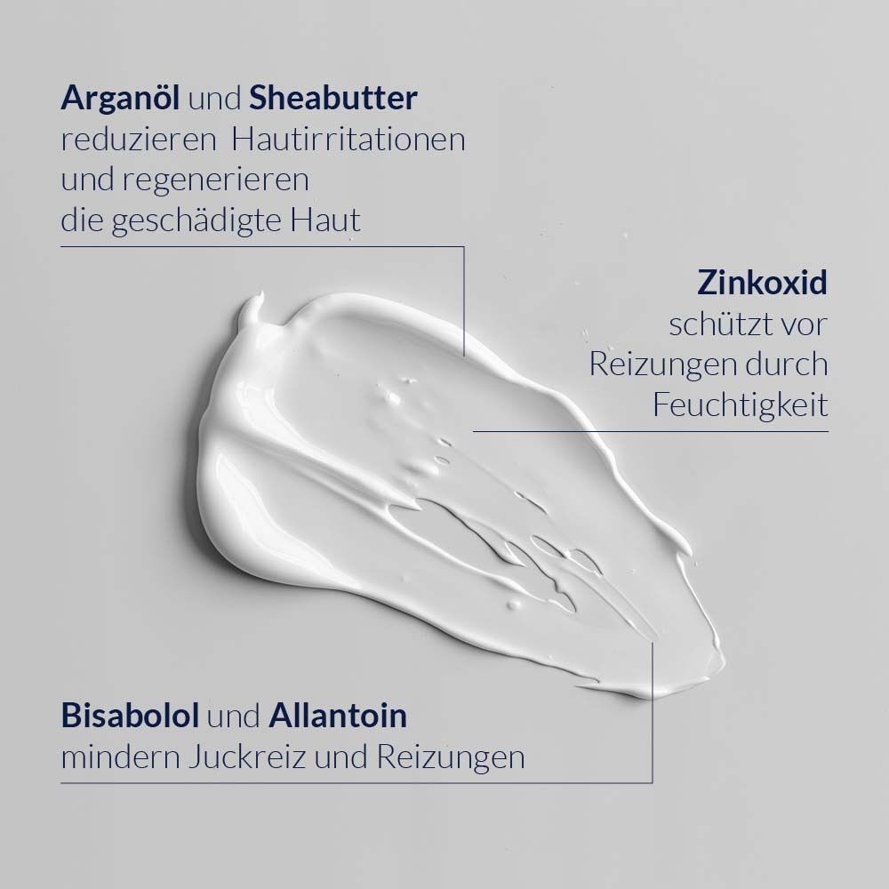 Nahaufnahme einer weißen Creme. Beschriftungen: Arganöl und Sheabutter, Zinkoxid, Bisabolol und Allantoin. Grauer Hintergrund.