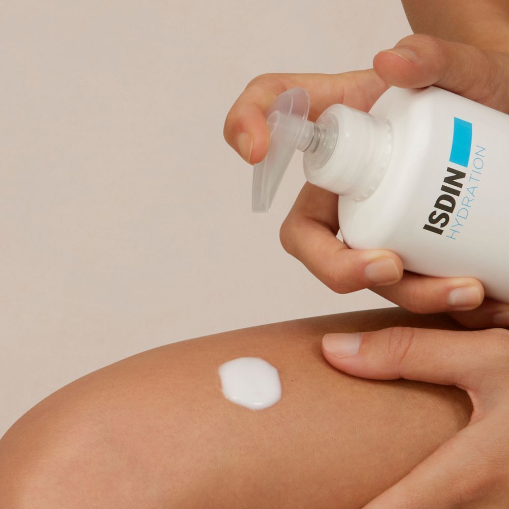 Hand drückt Lotion aus einer weißen Flasche mit blauem Etikett. Lotion wird auf Bein aufgetragen. Aufschrift: ISDIN Hydration.