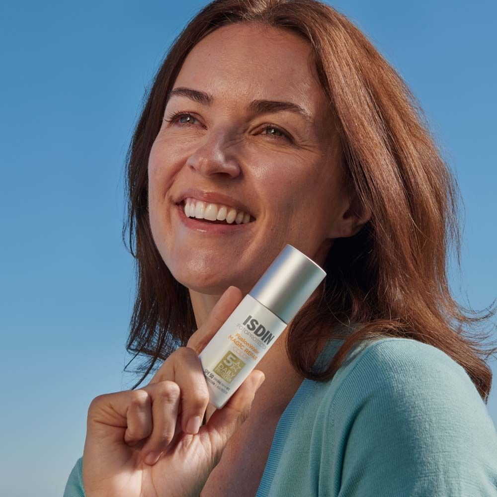Frau hält eine ISDIN Fotoprotector Fusion Water Magic Repair Color Flasche. Sie lächelt vor blauem Himmel.