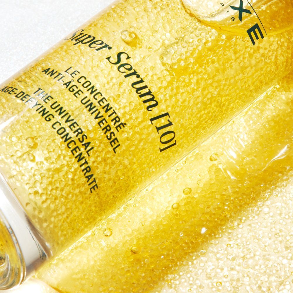 Nahaufnahme von NUXE Super Serum [10]. Goldfarbene Flüssigkeit mit sichtbaren Partikeln. Produktbeschriftung.