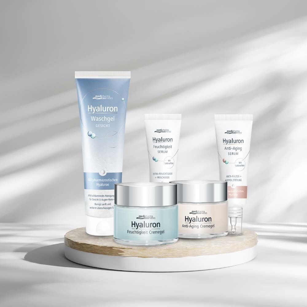 Verschiedene Produkte: Waschgel, Serum, Creme. Weiße Tuben und Tiegel mit blauer und weißer Beschriftung. Auf Holzpodest.