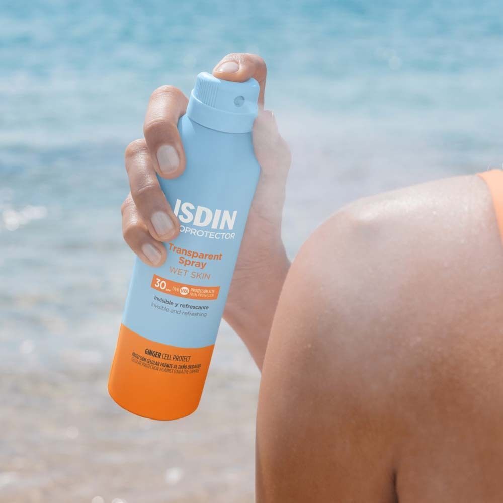Person sprüht mit einer blauen Flasche mit orangefarbenen Akzenten. Aufschrift: ISDIN Fotoprotector Transparent Spray Wet Skin 30.