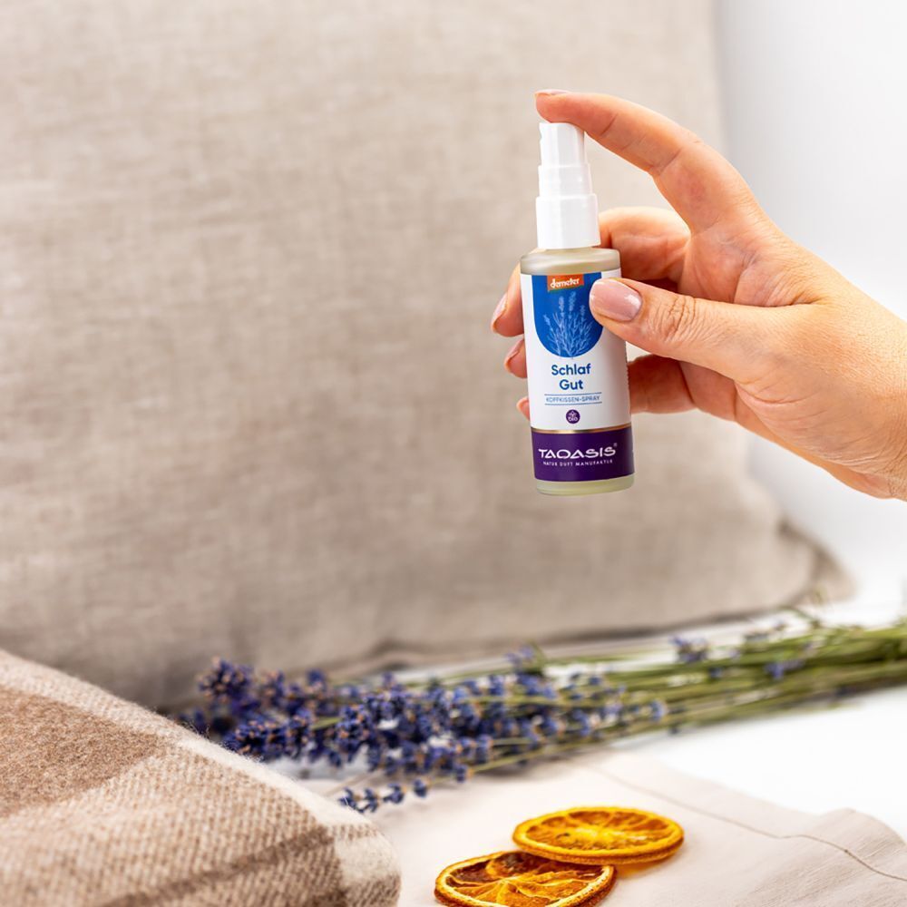 Hand hält eine Sprühflasche. Aufschrift: Schlaf Gut. Lavendel, Kissen und Decke im Hintergrund.