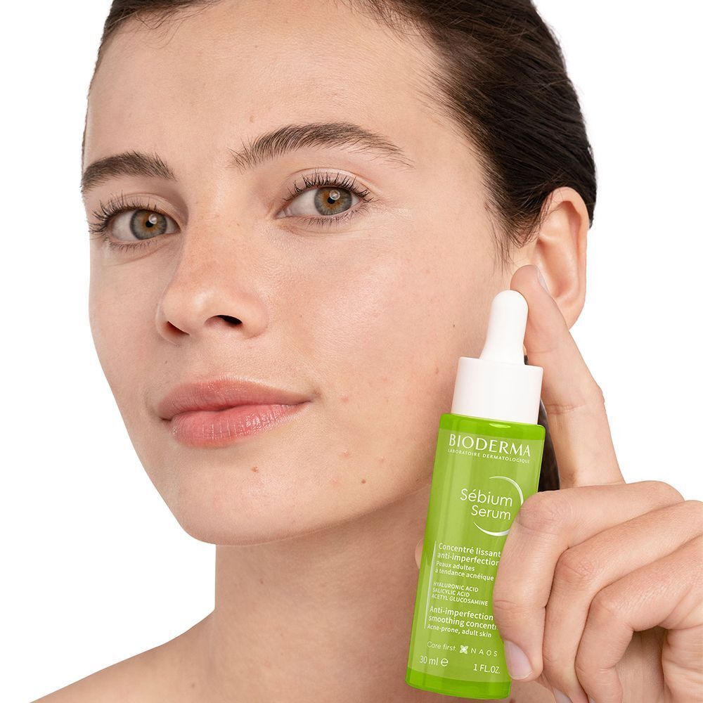 Frau hält eine grüne Flasche mit weißem Deckel. Aufschrift: BIODERMA, Sébium Serum. Anti-Unreinheiten, glättendes Konzentrat.