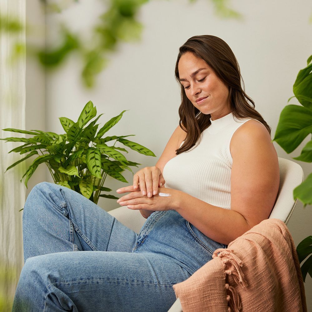 Frau sitzt in einem Stuhl und trägt Jeans und ein weißes Oberteil. Sie trägt Handcreme auf.