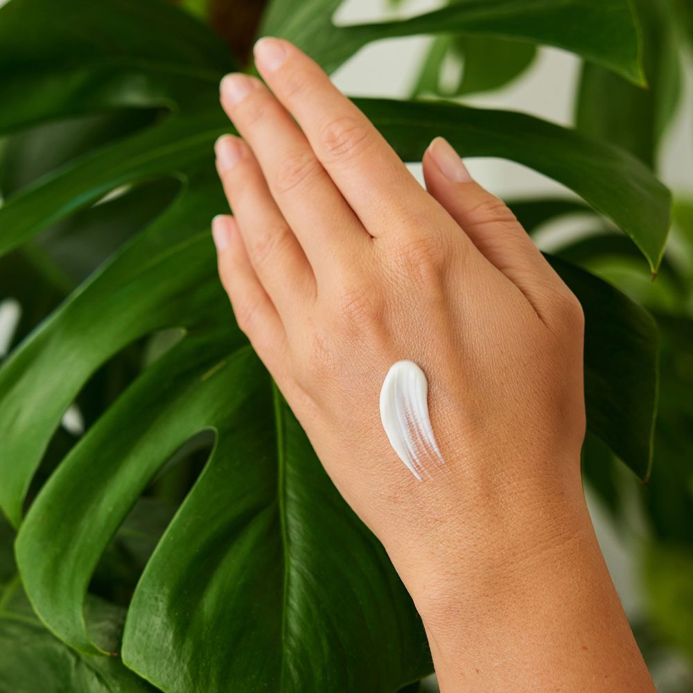 Hand mit einem Streifen weißer Creme auf der Handfläche, vor grünem Blattwerk.