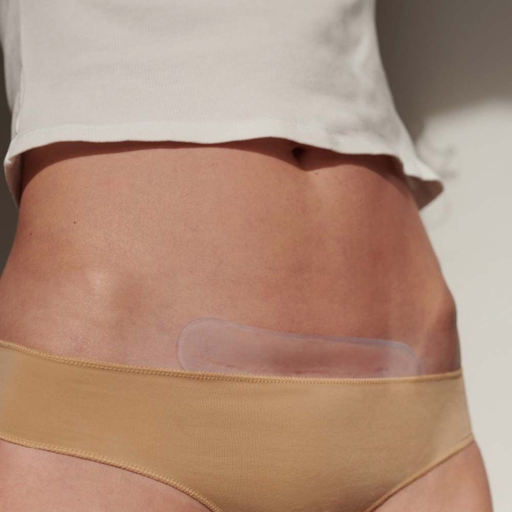 Bauch einer Person mit einem transparenten Pflaster auf der Haut. Person trägt Unterwäsche und ein weißes Oberteil.
