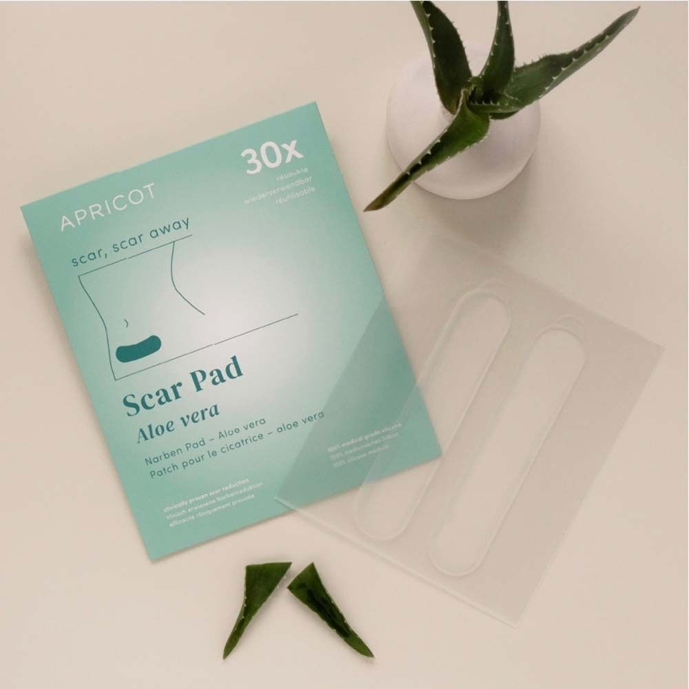 Verpackung und Pflaster neben Aloe Vera Blättern. Aufdruck: APRICOT, Scar Pad Aloe vera. Text: 30x.