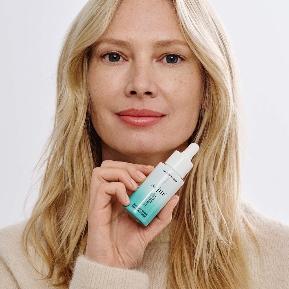 Frau mit blonden Haaren hält eine Flasche Anti-Pigment Serum. Die Flasche ist weiß mit grünem Deckel und Schriftzug.