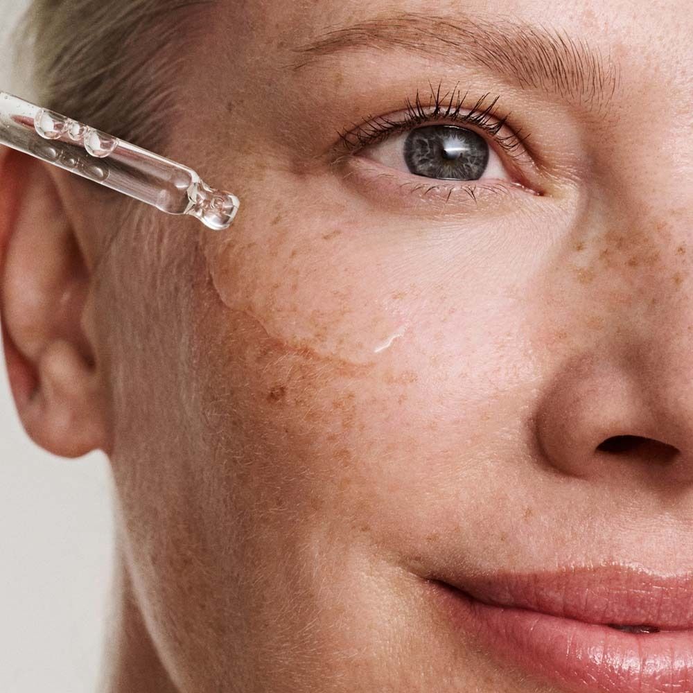 Nahaufnahme eines Gesichts mit einem Tropfen Anti-Pigment Serum. Die Haut zeigt Sommersprossen und das Serum wird aufgetragen.