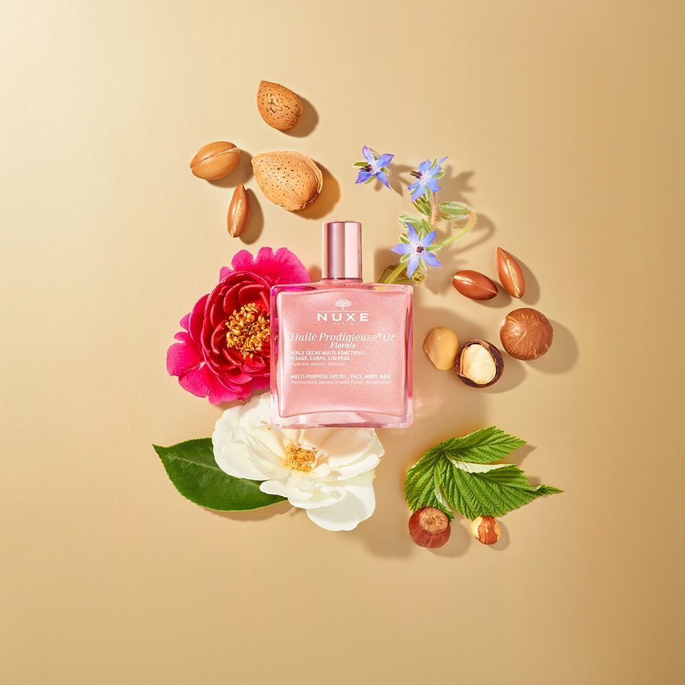 Ein Flakon mit rosafarbenem Öl, umgeben von Blüten und Nüssen. Aufschrift: Nuxe Huile Prodigieuse® Or Florale. Quadratischer Flakon mit goldenem Verschluss.