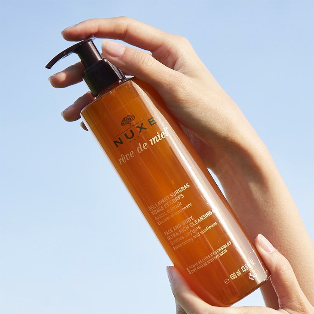 Hand hält eine Flasche NUXE Rêve de Miel® Duschgel. Orangefarbene Flasche mit braunem Pumpaufsatz.