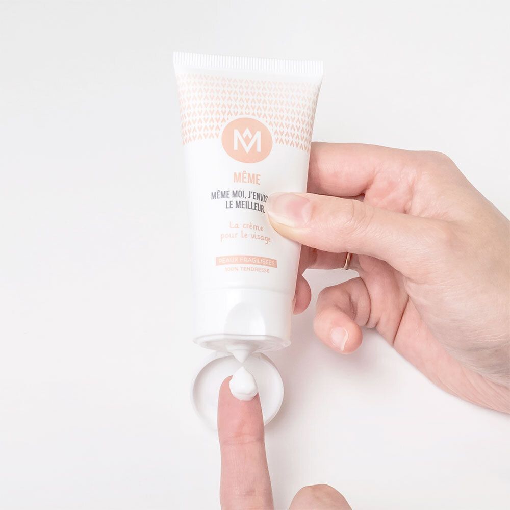 Hand hält Tube MÊME Gesichtscreme. Creme wird auf Finger und Unterlage gedrückt. Aufschrift: MÊME, MÊME MOI, J'ENVISAGE LE MEILLEUR. La crème pour le visage. Peaux fragilisées.