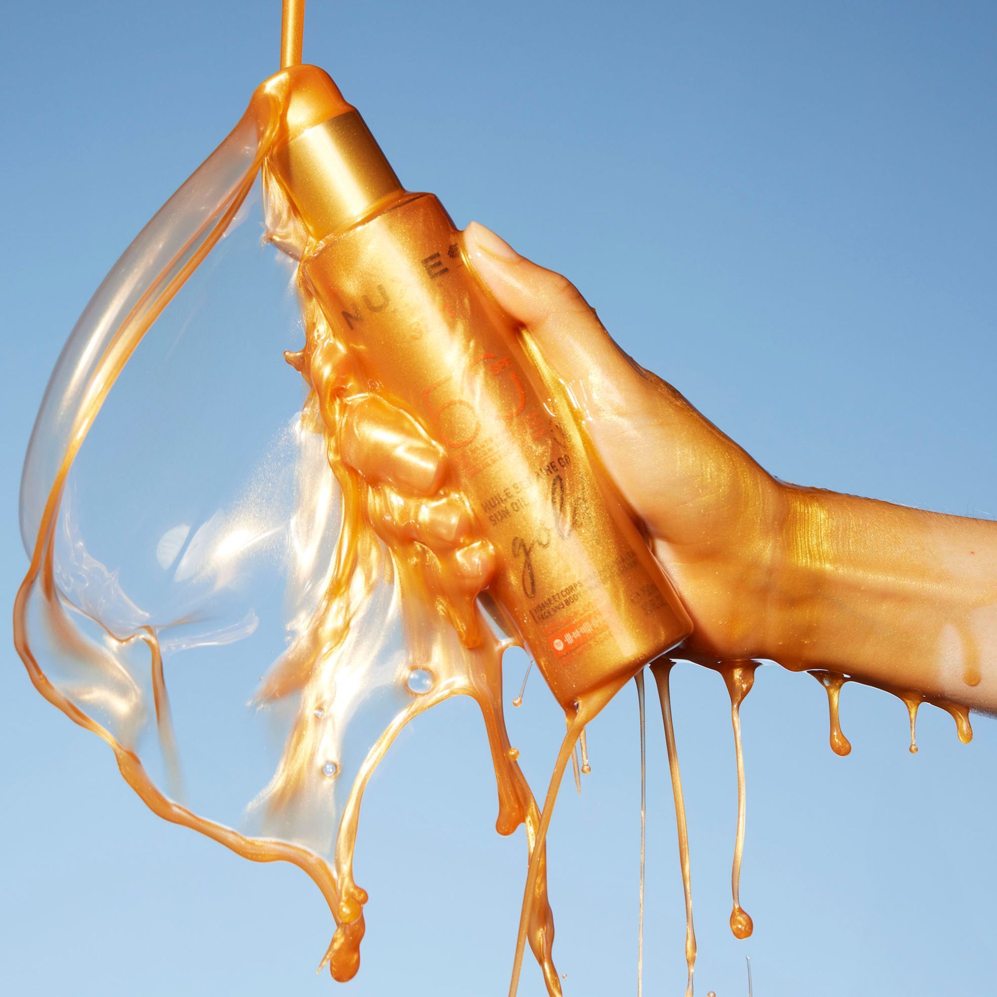 Hand hält eine goldfarbene Sonnenölflasche. Goldfarbene Flüssigkeit fließt aus der Flasche.