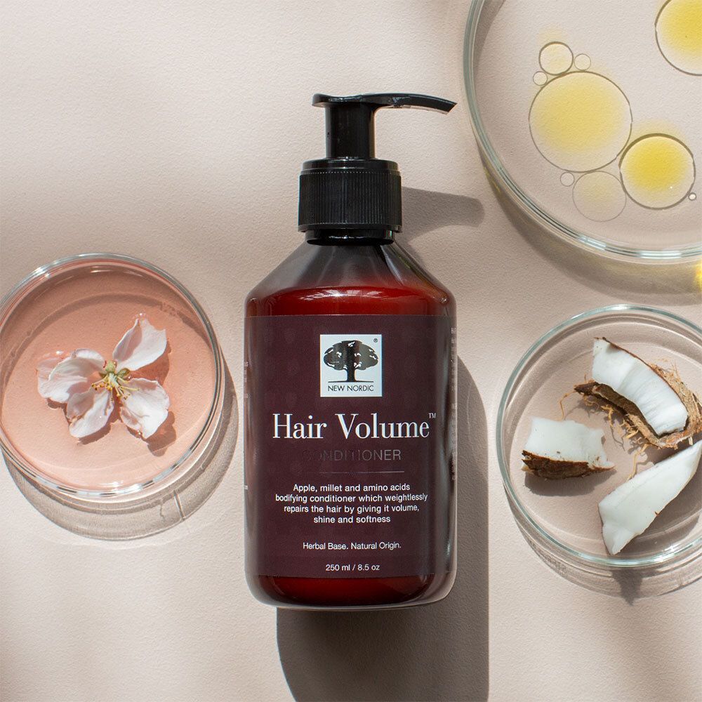 Flasche NEW NORDIC Hair Volume™ Conditioner, umgeben von Blüten und Schalen mit Inhaltsstoffen. Braune Flasche mit schwarzem Pumpverschluss.