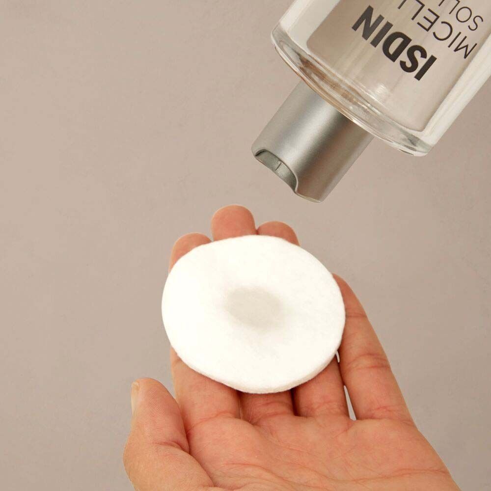 Eine Hand hält ein Wattepad, das mit ISDIN Mizellenwasser befeuchtet wird. Die Flasche ist im Hintergrund zu sehen.