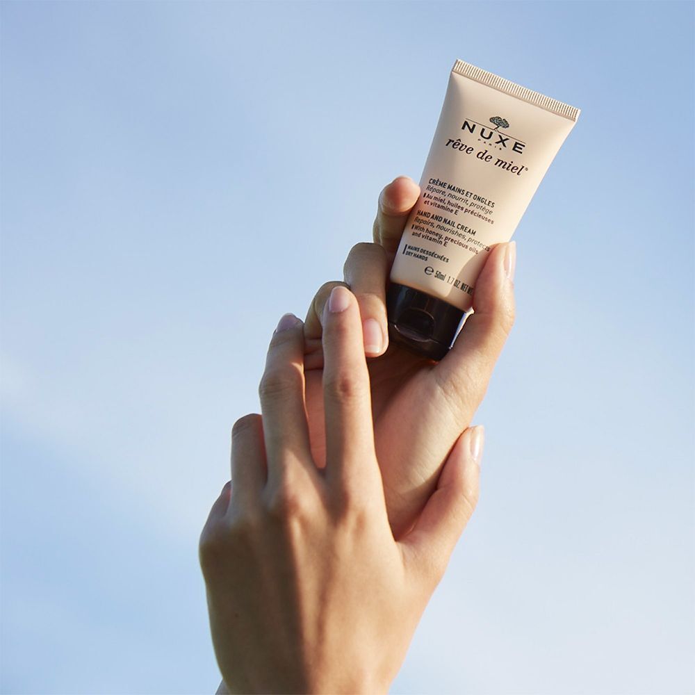 Hände halten eine Tube NUXE Rêve de Miel® Hand- & Nagelcreme. Himmelblauer Hintergrund. Produkt im Fokus.