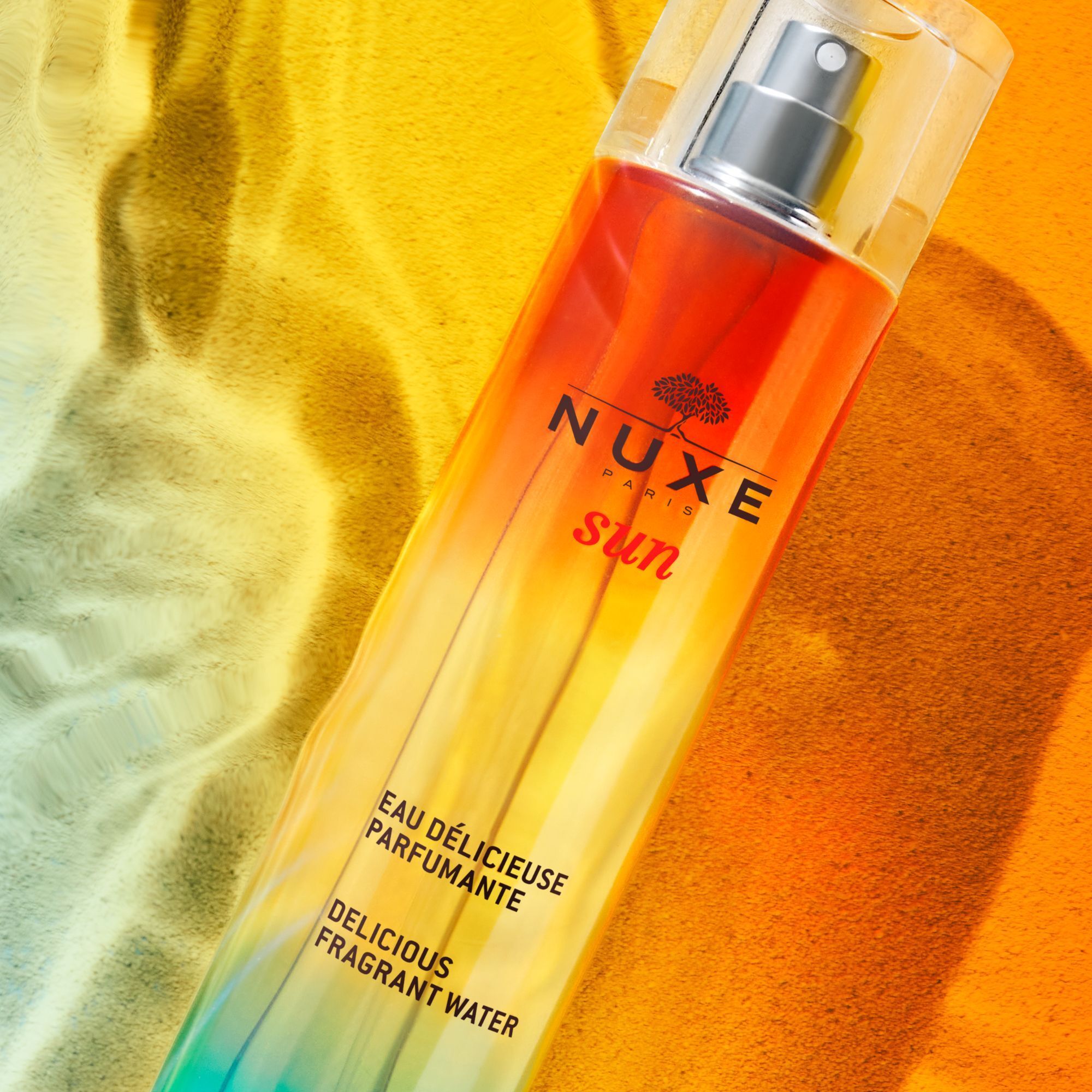 NUXE Sun Eau Délicieuse Parfumante. Flakon auf sandfarbenem Hintergrund. Produktname und Logo sichtbar.