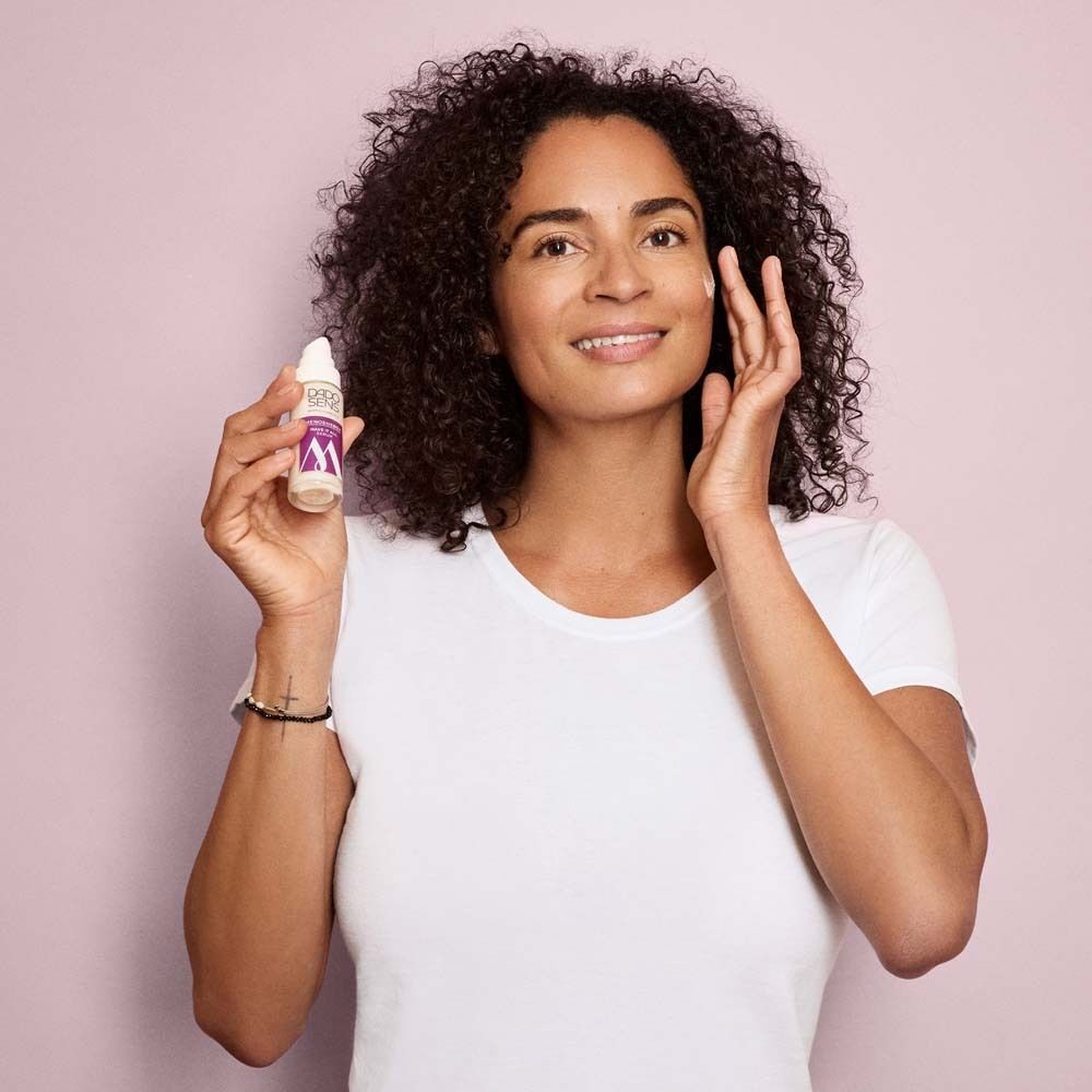 Frau hält eine Flasche DADO SENS Menoenergy Serum. Sie berührt ihr Gesicht. Hintergrund: Rosa.