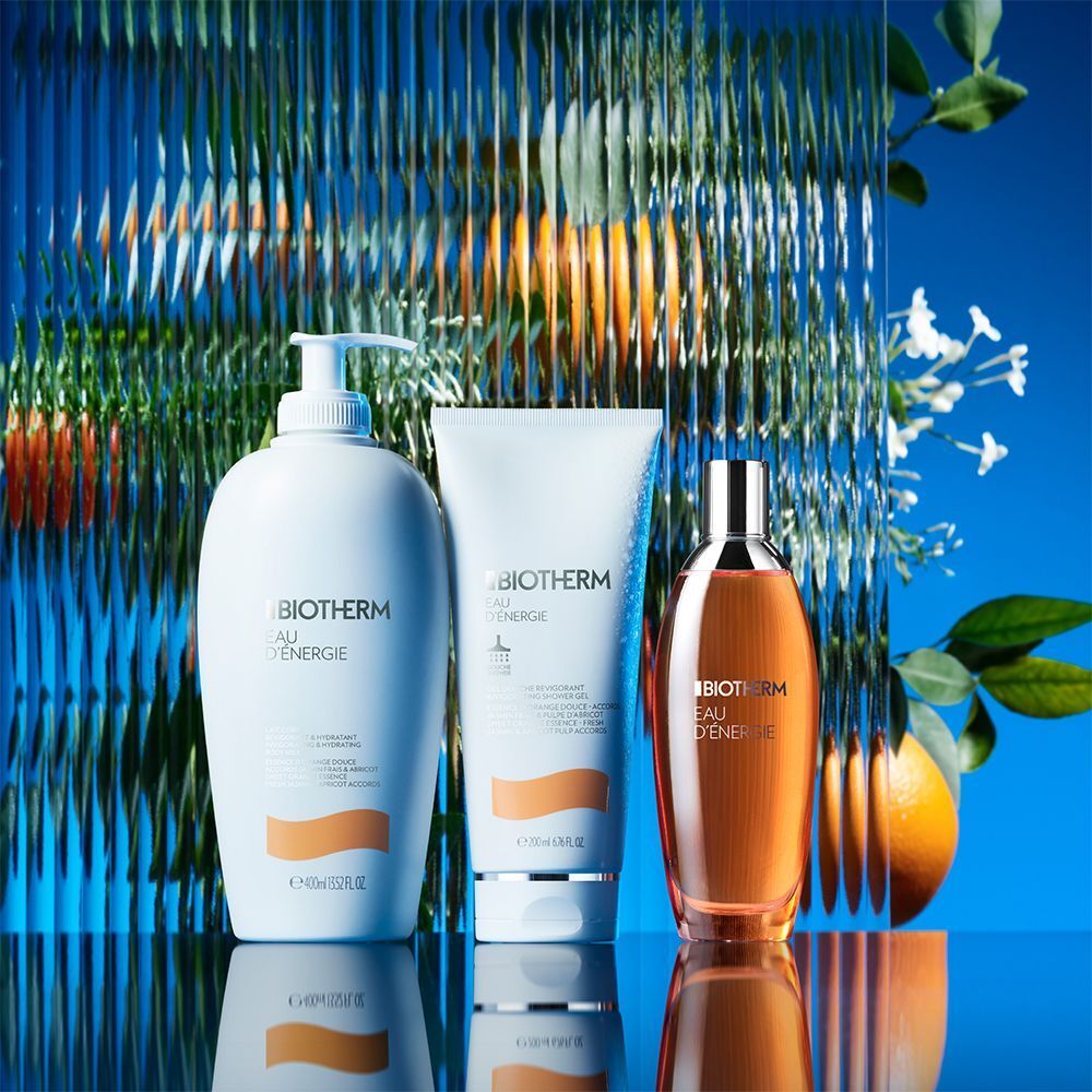 Drei BIOTHERM Eau d'Energie Produkte: Flasche, Tube und Spray. Weiß, orange und transparent. Text: BIOTHERM, Eau d'Energie. Hintergrund: blau und grün.