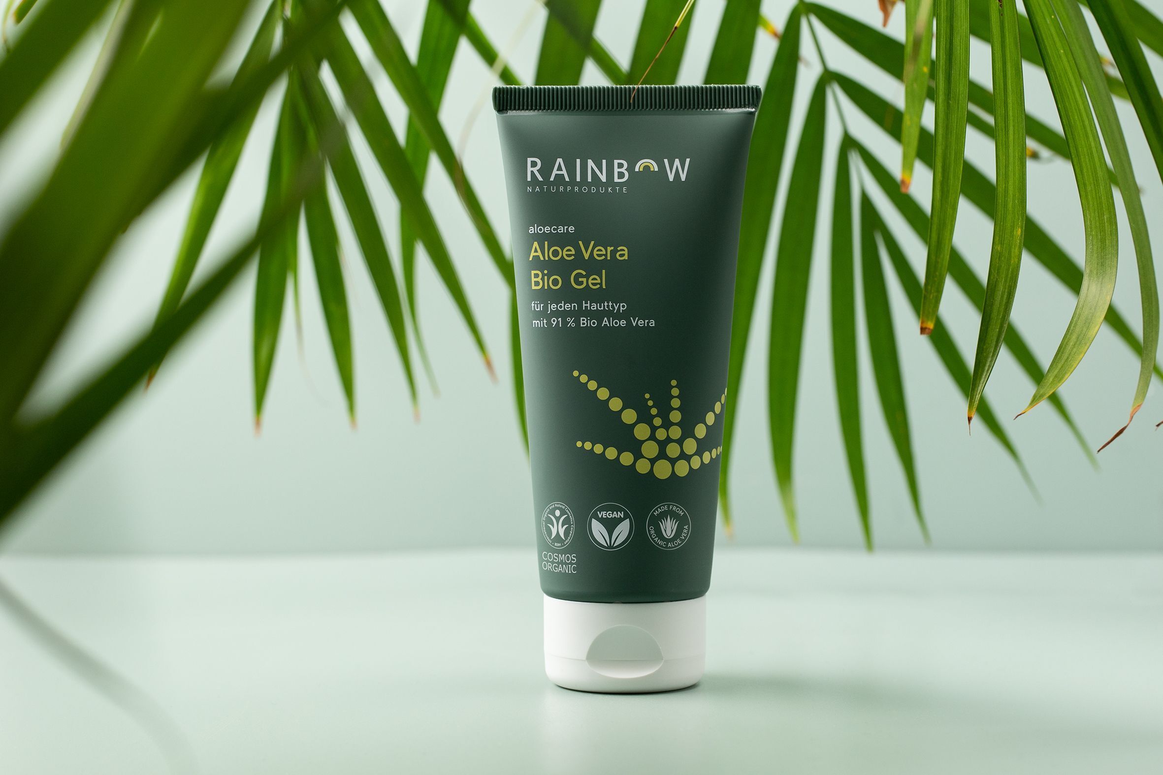 Grüne Tube mit weißem Verschluss. Aufschrift: RAINBOW Naturprodukte, Aloe Vera Bio Gel, 91% Bio Aloe Vera, für jeden Hauttyp. Logos. Palmblätter.