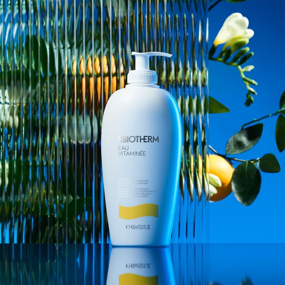 Weiße Flasche BIOTHERM Eau Vitaminée Bodymilk mit Pumpverschluss. Gelbes Etikett. Hintergrund: blau, Spiegelung, Pflanzen.