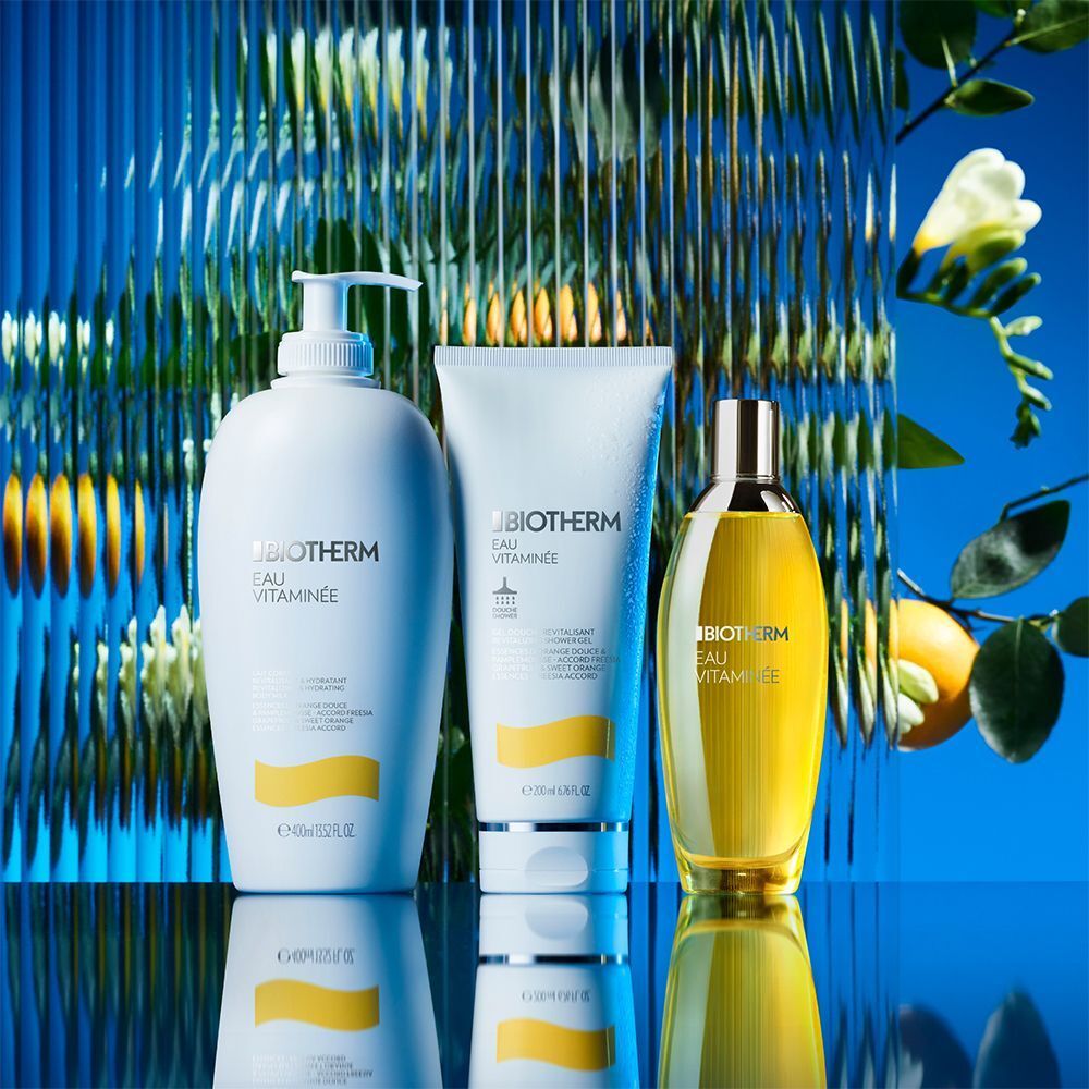 Drei Produkte: BIOTHERM Eau Vitaminée Bodymilk, Duschgel und Bodyspray. Hintergrund: blau, Spiegelung, Pflanzen.