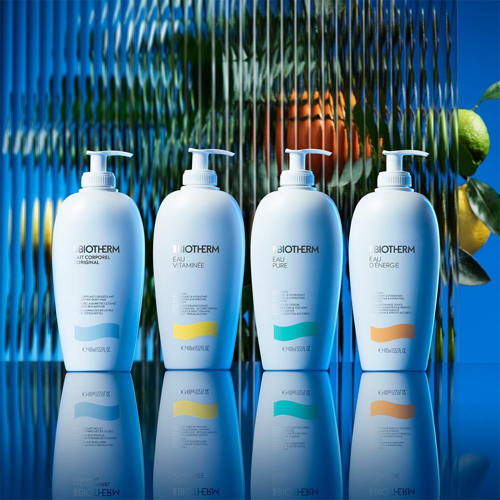 Vier weiße Flaschen BIOTHERM Eau Vitaminée Bodymilk mit Pumpverschluss. Verschiedene Etikettenfarben. Hintergrund: blau, Spiegelung.