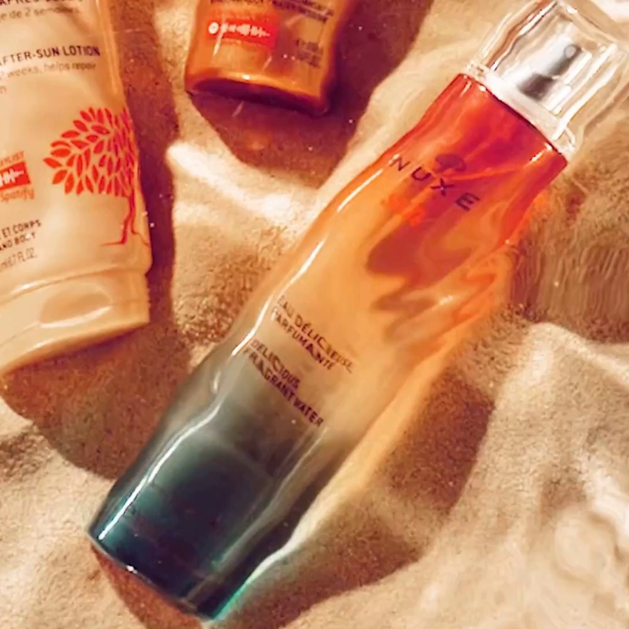 Flakon mit Sprühkopf, daneben eine Tube und eine weitere Flasche. Auf Sand. Text: NUXE Sun, Eau Délicieuse Parfumante.