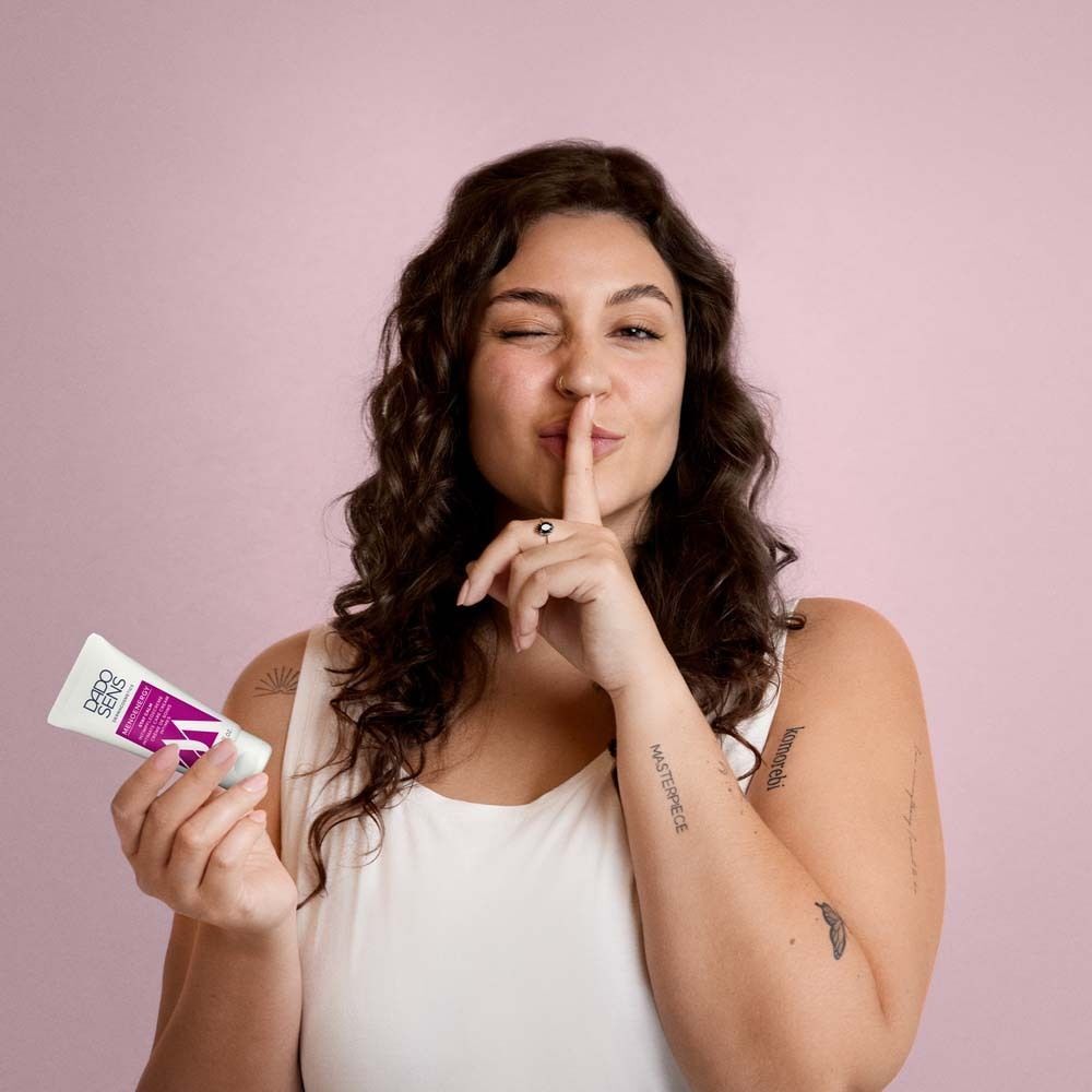 Frau mit DADOSENS MENOENERGY SLEEP WELL Nachtcreme-Tube. Sie hält Finger an Lippen, zwinkert. Hintergrund rosa.