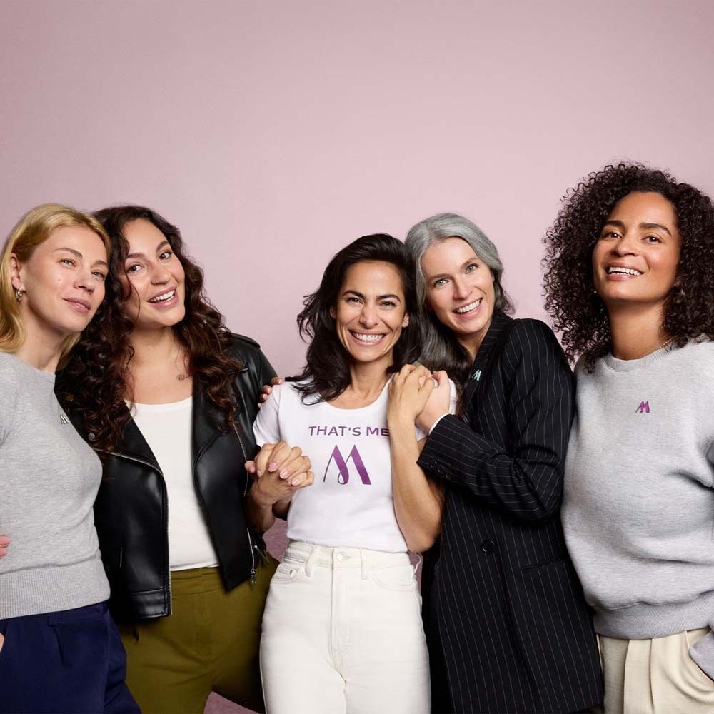 Fünf Frauen stehen zusammen. Sie tragen Shirts mit Logo. Hintergrund rosa.