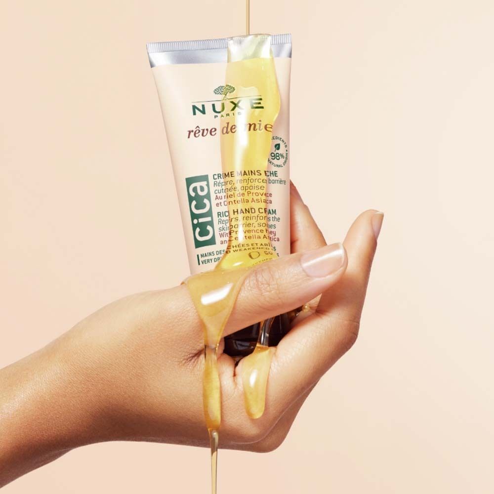 Hand hält eine Tube Handcreme. Creme fließt aus der Tube und über die Hand. Aufdruck: Nuxe, Rêve de Miel, Cica.