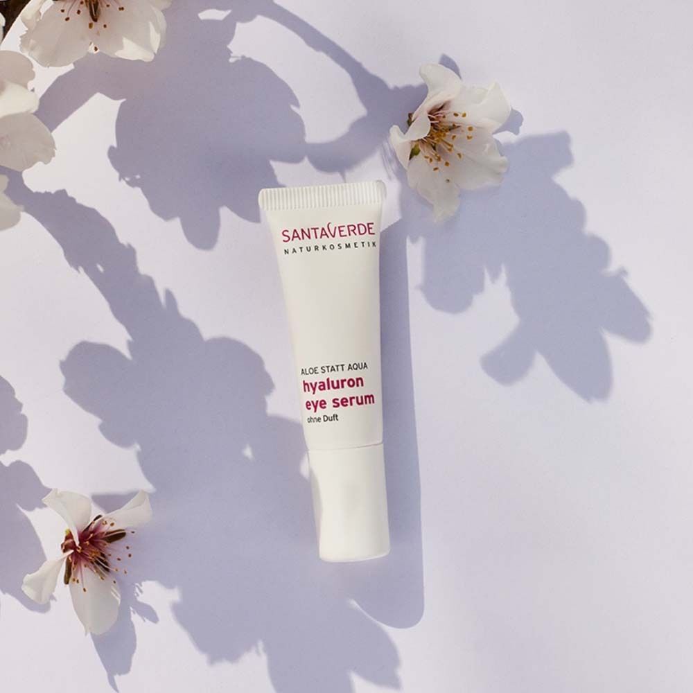 Eine Tube SANTAVERDE Hyaluron Augenserum. Weiße Tube mit lila Schriftzug. Aufschrift: FACE CARE, hyaluron eye serum, ohne Duft. Dekoriert mit Blüten.