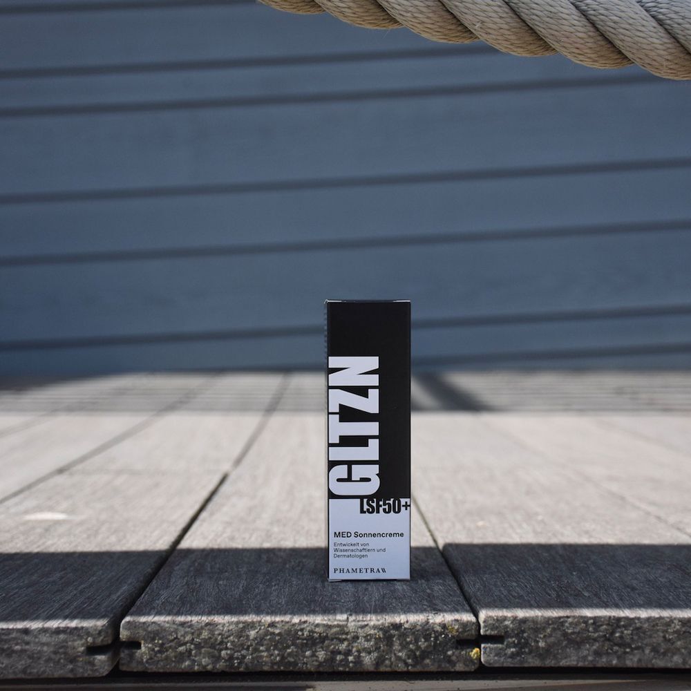 Eine Schachtel Sonnencreme steht auf einem Holzsteg. Auf der Schachtel steht GLTZN LSF 50+. MED Sonnencreme. PHAMETRA.