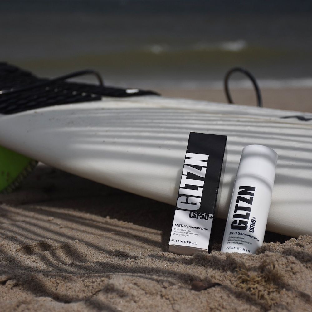 Eine Tube und eine Schachtel Sonnencreme am Strand. Auf der Tube und Schachtel steht GLTZN LSF 50+. MED Sonnencreme. PHAMETRA.