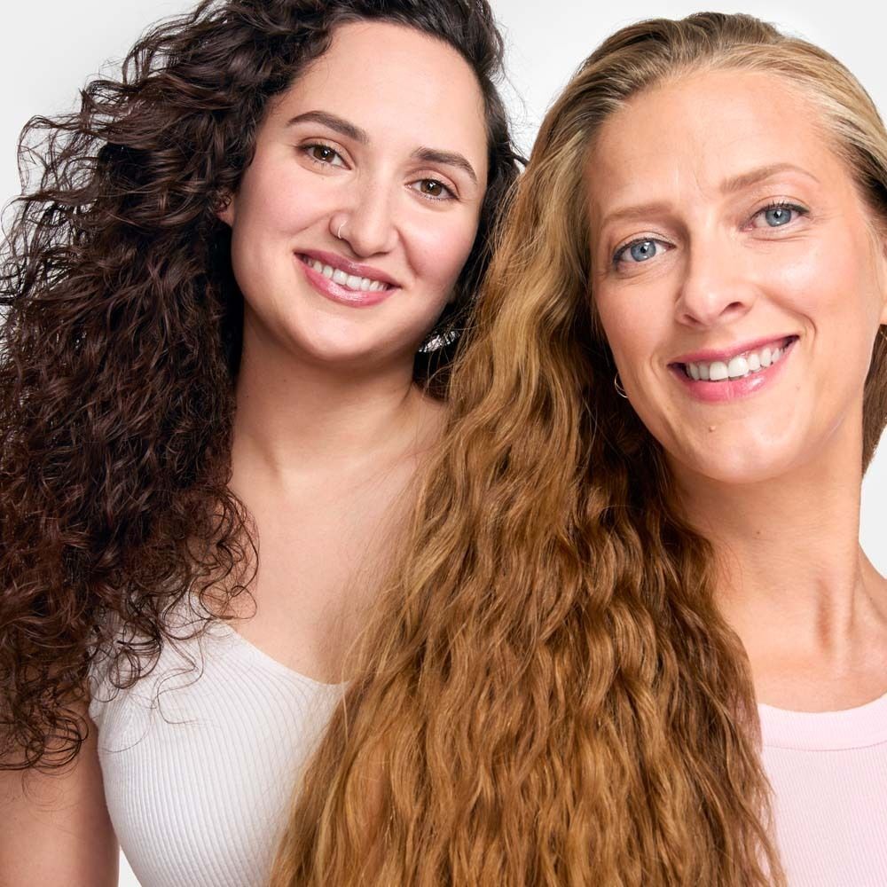 Zwei Frauen mit lockigem und welligem Haar. Eine Frau hat braunes Haar, die andere blondes Haar. Beide lächeln.