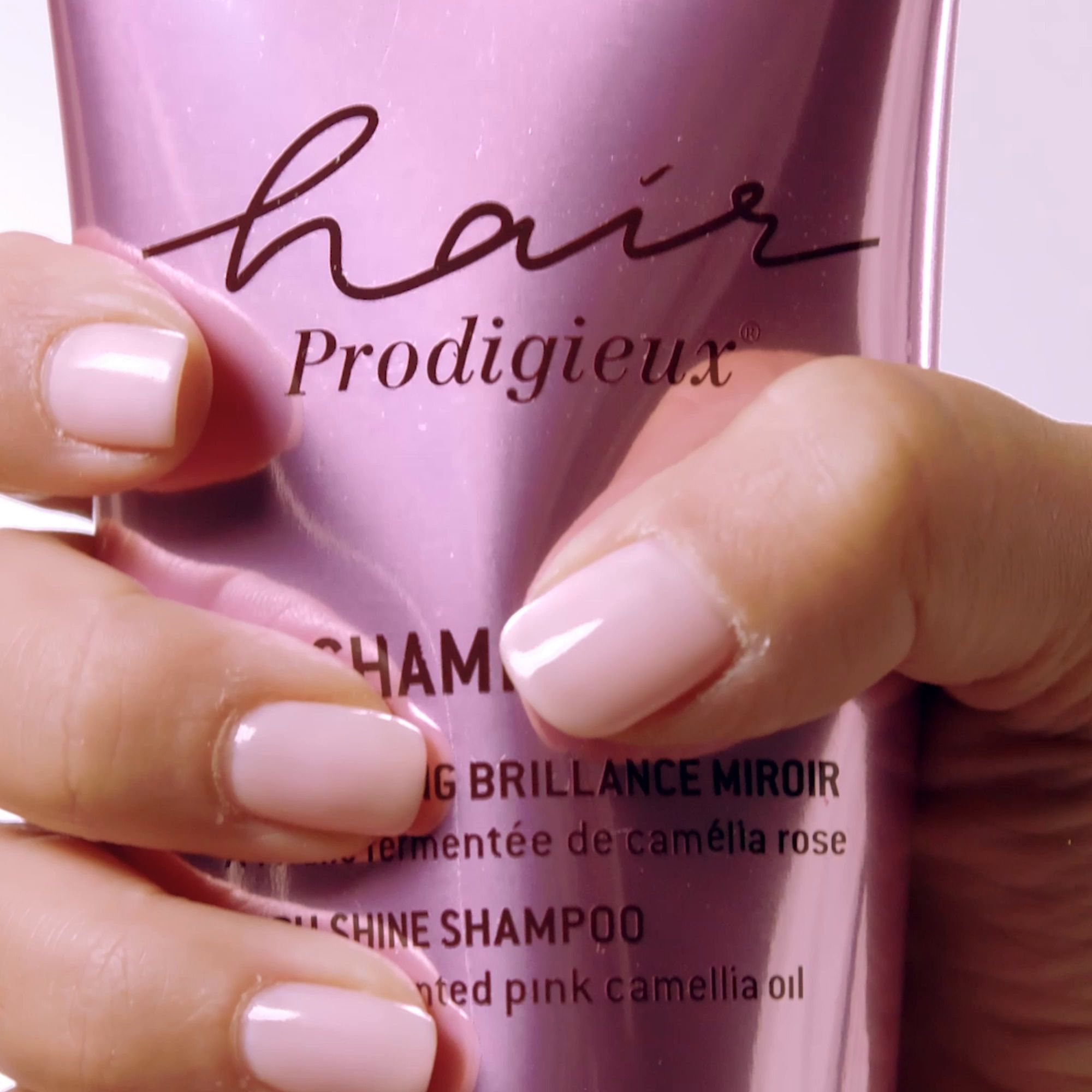 Nahaufnahme einer Hand, die eine rosa Tube Shampoo hält. Aufschrift: Hair Prodigieux, Shampooing Brillance Miroir, High Shine Shampoo.