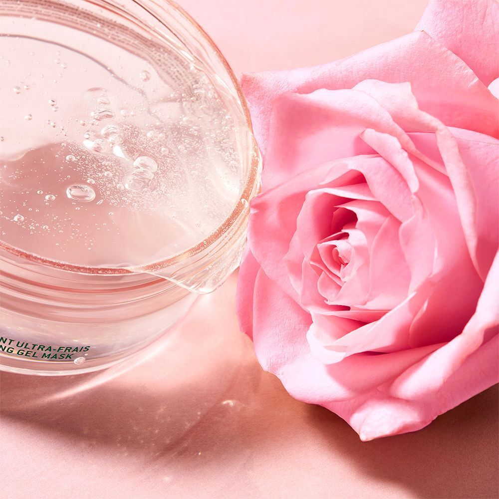 Durchsichtige Flüssigkeit in einem Glasbehälter, daneben eine rosa Rose. Flüssigkeit mit Bläschen. Hintergrund rosa.