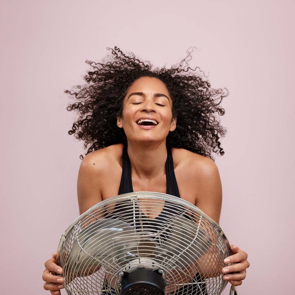 Eine Frau mit lockigem Haar steht vor einem Ventilator. Sie lächelt und hat die Augen geschlossen.