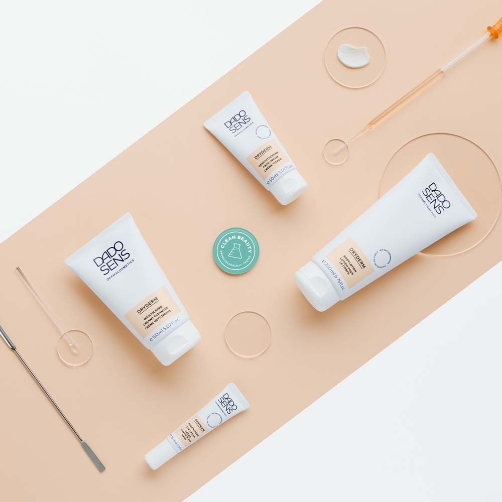 Verschiedene DADO SENS Produkte auf beigem Hintergrund. Tuben, Tiegel, Pipette, Spatel und ein grünes Siegel.