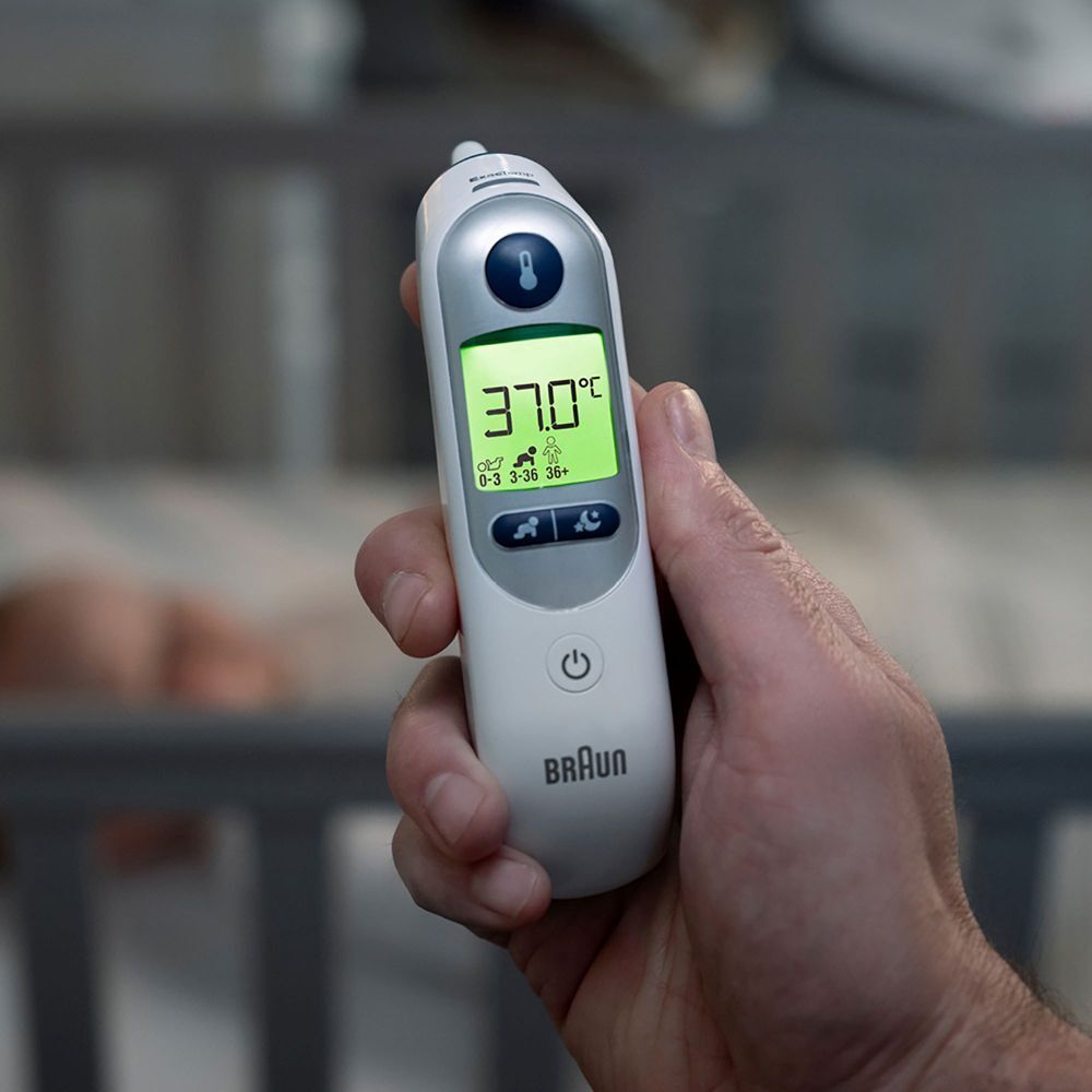 Hand hält Braun ThermoScan 7+. Display zeigt Temperatur. Hintergrund: Babybett.