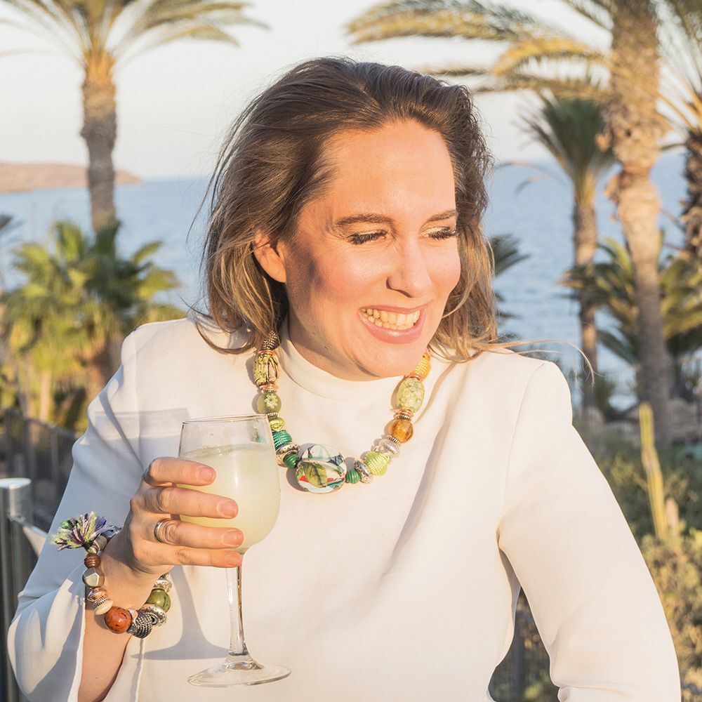 Frau mit Glas in der Hand, lächelnd. Sie trägt Schmuck und ein weißes Oberteil. Hintergrund: Meer und Gebäude.