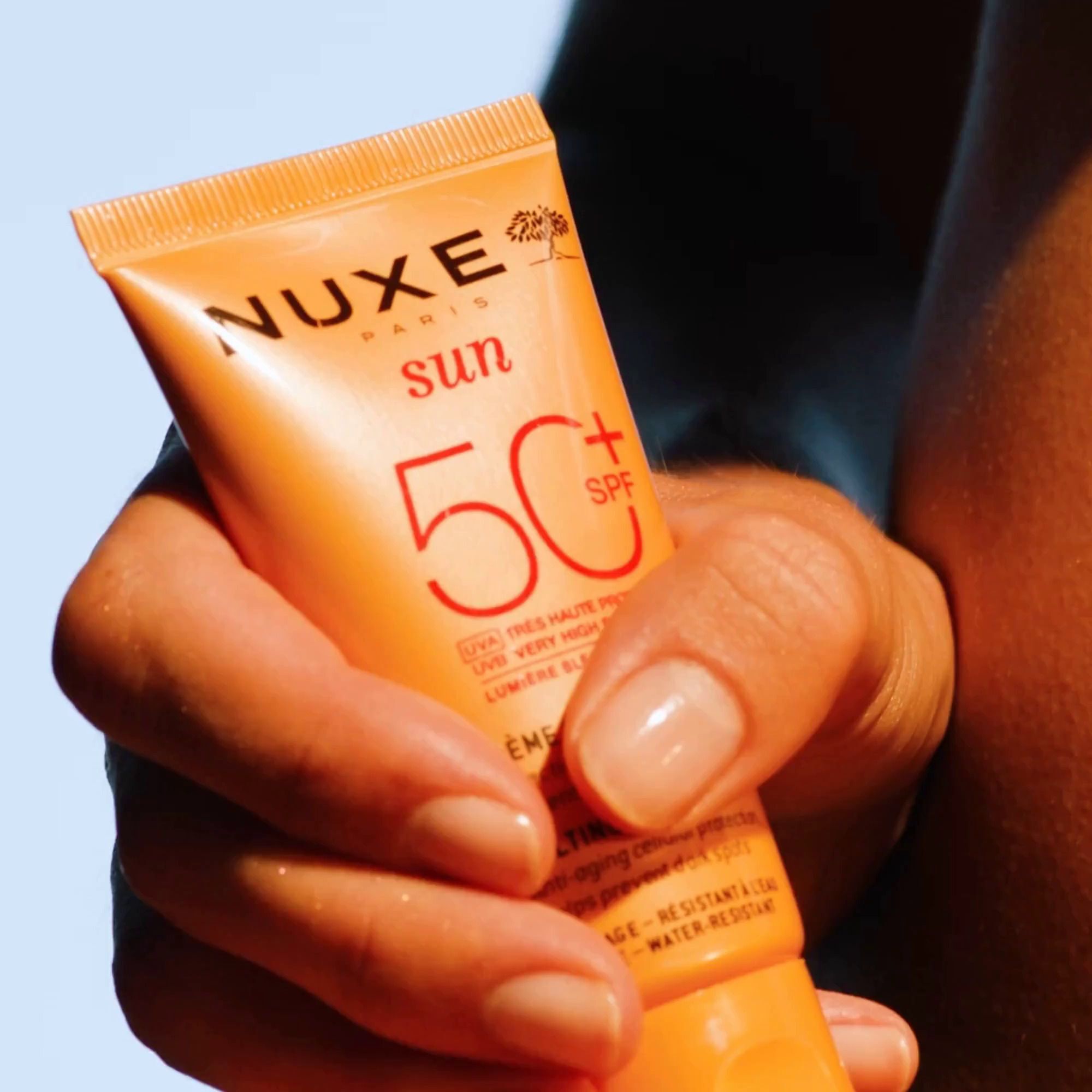 Hand hält eine Tube Nuxe Sun Sonnencreme Gesicht LSF 50, orangefarben.