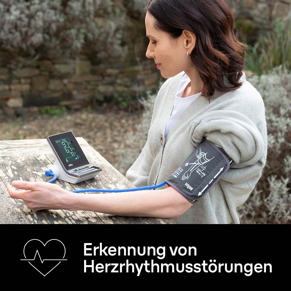 Frau misst Blutdruck mit Gerät und Manschette. Gerät mit Display. Manschette am Arm. Hintergrund: Garten.