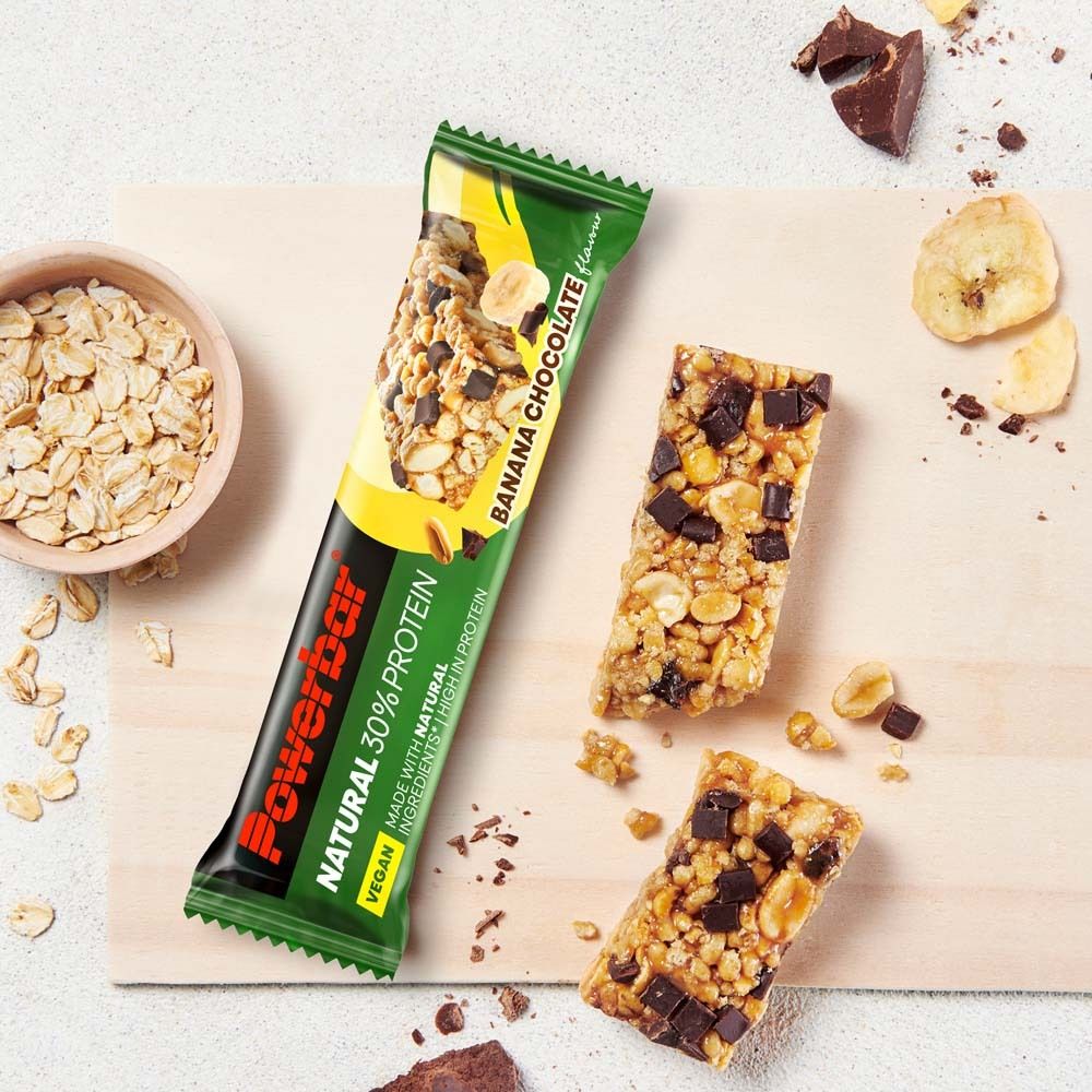 Powerbar NATURAL PROTEIN Banane-Schokolade Riegel und Stücke. Auf Holzbrett. Grüne Verpackung. Text: 30% Protein, vegan.