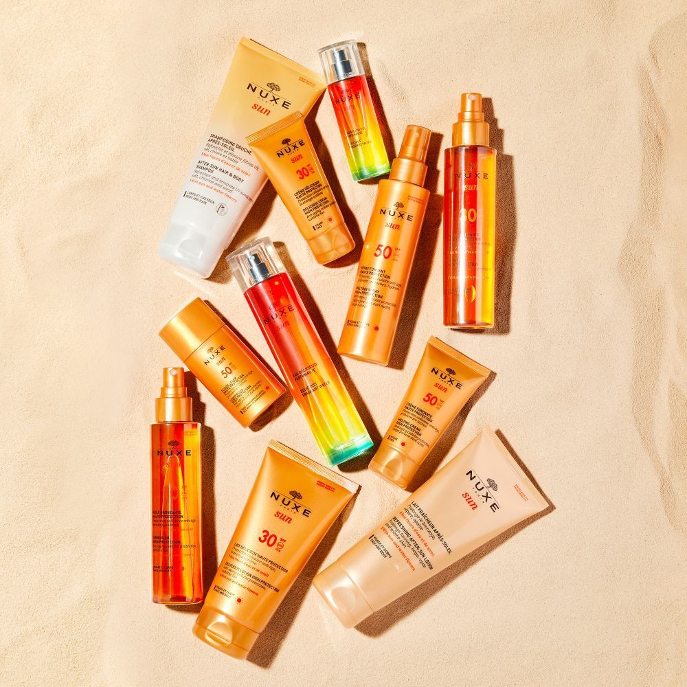 Verschiedene NUXE Sun Produkte auf sandigem Untergrund. Flaschen und Tuben in Gold- und Orangetönen.