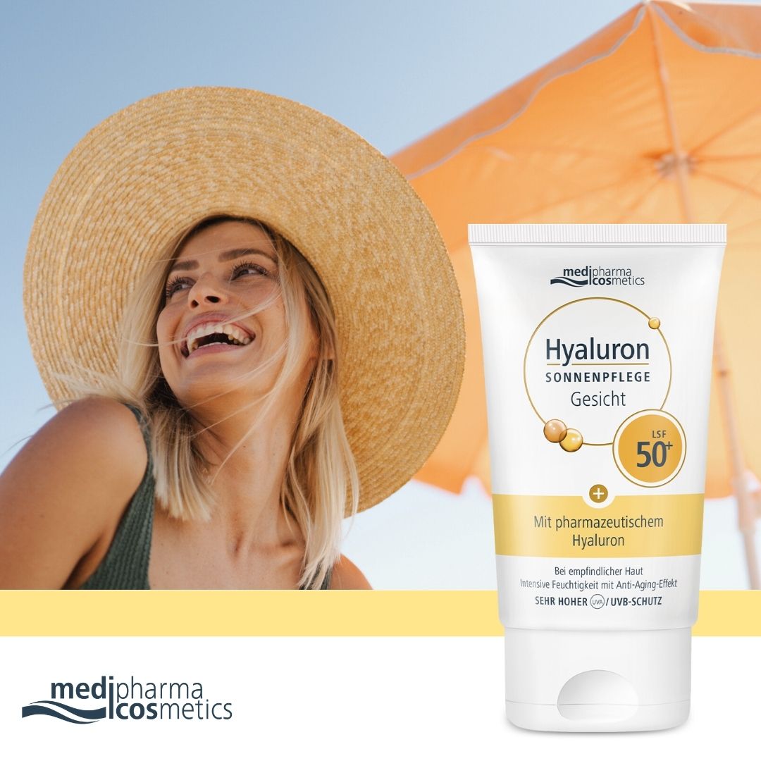 Frau mit Hut und Sonnencreme-Tube. Tube: medipharma cosmetics Hyaluron Sonnenpflege Gesicht LSF 50+. Hintergrund: Strand und Sonnenschirm.