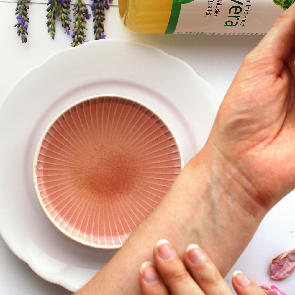 Arm mit aufgetragener Flüssigkeit auf einer rosa Platte. Im Hintergrund eine Flasche Aloe vera Gel-Saft.