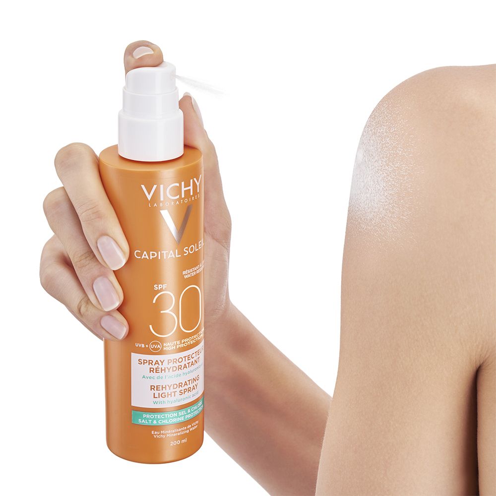 Hand hält eine orangefarbene Sonnencreme-Flasche. Sprühkopf zeigt auf einen Arm. Aufschrift: VICHY, CAPITAL SOLEIL, SPF 30.