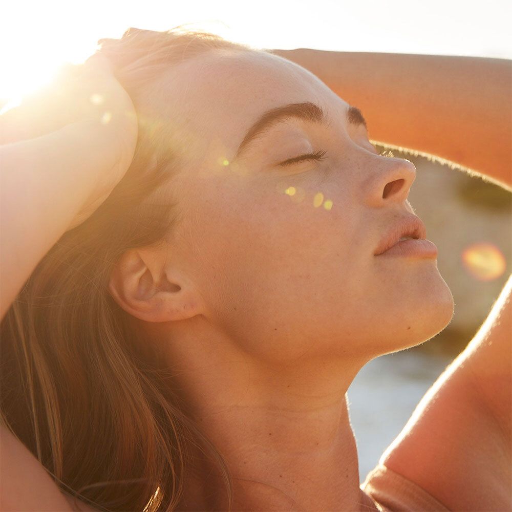 Eine Frau mit geschlossenen Augen, die ihr Gesicht in die Sonne hält. Ihr Haar ist leicht gewellt.